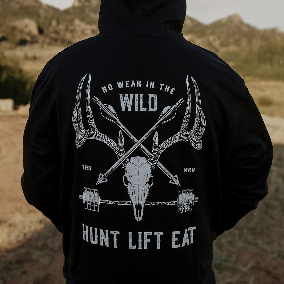A person standing outdoors in a desert landscape, viewed from behind, wearing a black hoodie with a large white graphic on the back featuring antlers, a skull, crossed arrows, and the text 'NO WEAR IN THE WILD' and 'HUNT LIFT EAT'.